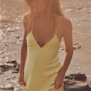 Ljusgul långklänning i satin - Säljer en ljusgul långklänning med smala axelband och v-ringning. Klänningen har en snygg öppen rygg med dekorativa band och är tillverkad i glansig satin som ger en lyxig känsla. Perfekt för dig som vill sticka ut med en enkel men elegant look.