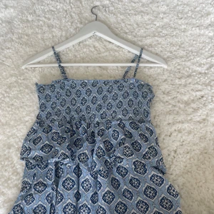 Blå mönstrad sommartopp Gina Tricot - Supersöt blå topp från Gina Tricot med smala axelband och ett bohemiskt mönster i vitt och mörkblått. Toppen har en smockad överdel och volangdetaljer, perfekt för varma dagar. Lätt och luftig i bomullsmaterial.