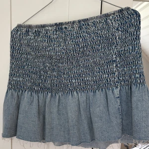 Smockad jeans topp med volang - Supersöt ljusblå jeans topp  med smockad midja och bred volang nedtill. Toppen har en peplum-effekt och är gjord i mjukt denimtyg. Perfekt att styla med en jeansjacka för en chill look. Passar dig som gillar trendiga och bekväma plagg. Använd två gånger