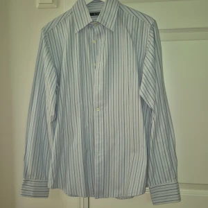 Randig skjorta från Tiger of Sweden - Snygg skjorta från Tiger of Sweden i storlek 40. Skjortan är vit med tunna blå ränder, har klassisk krage och långa ärmar med dubbla knappar vid manschetten. Tillverkad i bomull och har en normal passform. Perfekt för dig som gillar stilrena och fräscha plagg.