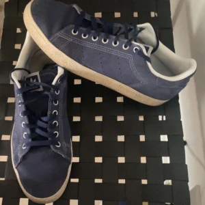 Adidas Stan Smith blå mocka sneakers - Säljer ett par Adidas Stan Smith sneakers i mörkblå mocka med vita detaljer och klassisk platt sula. Skorna har snörning och rund tå, samt diskreta perforeringar på sidorna. Perfekt för dig som gillar stilrena och ikoniska sneakers med en twist.