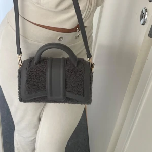 Handväska i teddy och skinn (Grå)  - Säljer en snygg grå handväska med både axelrem och handtag. Väskan har en unik design med mjukt teddy-material och detaljer i grått skinn. Perfekt storlek för det viktigaste och har fina guldfärgade metalldetaljer.