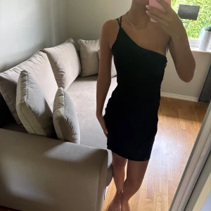 Svart one shoulder kortklänning - Säljer en svart kortklänning med one shoulder-design och smala axelband. Klänningen har en tight passform och är perfekt för dig som gillar en stilren och enkel look. Materialet känns mjukt och följsamt mot kroppen.
