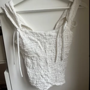 Vit linnetopp med knytband - Supersöt vit linnetopp med breda axelband som knyts på axlarna. Toppen har en krinklad struktur och är ärmlös, perfekt för varma dagar. Enkel och stilren design som passar till jeans eller kjol.