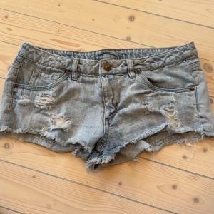 Blå jeansshorts med slitningar - Säljer ett par grå jeansshorts med coola slitningar och fransiga kanter. Perfekta för en avslappnad stil. De har en knapp och dragkedja framtill samt fickor både fram och bak.