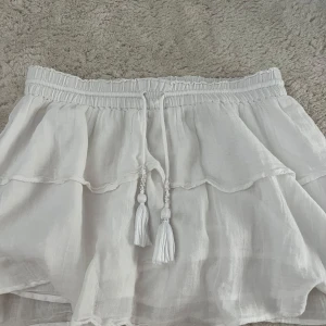 Vit kortkjol med volang från Gina Tricot - Supersöt vit kortkjol från Gina Tricot med volang och dekorativa snören med pärlor och tofsar i midjan. Kjolen har en elastisk midja och är gjord i ett lätt och luftigt linnematerial, perfekt för varma dagar. Passformen är normal och kjolen har en fräsch och ungdomlig vibe.