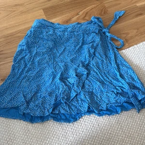 Blå omlottkjol med volang - Supersöt blå omlottkjol med små vita blommor och volangkant. Kjolen har knytband i midjan och är perfekt för dig som gillar en feminin och chill stil. Lätt och skön att bära under varma dagar.