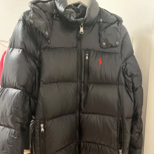Svart dunjacka från Polo Ralph Lauren - Snygg svart dunjacka från Polo Ralph Lauren med röd logga på bröstet. Finns små defekter som små hål men inget man märker när man använder och de syns inte heller. Köpt från ett känt företag här på Plick vinofino. Köpte den för 4000kr. Perfekt att köpa nu innan säsongen börjat då konkurrensen snart blir större plus högre priser