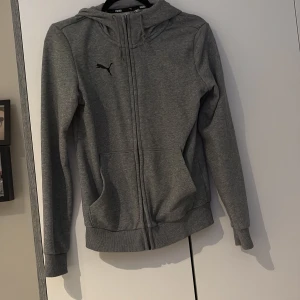 Grå hoodiejacka från Puma - Grå hoodiejacka från Puma med dragkedja framtill, huva med snörning och två stora fickor. Jackan har ribbade muddar vid ärmslut och nederkant samt svart Puma-logga på bröstet. Perfekt för en avslappnad och sportig stil.