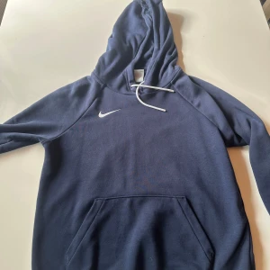 Marinblå Nike hoodie med huva - Säljer en marinblå hoodie från Nike i storlek S. Tröjan har en klassisk huva med vita snören, en stor magficka och Nike-logga på bröstet. Perfekt för chill dagar eller när du vill ha en sportig look. Materialet är mjukt och skönt, troligen bomull och polyester.