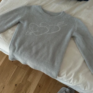Ljusgrå fluffig stickad tröja YAMZ - Supermysig ljusgrå stickad tröja från YAMZ med lång ärm och rund hals. Tröjan har ett fluffigt material och ett diskret hjärtmönster framtill. Perfekt för kalla dagar när du vill vara både comfy och cute.