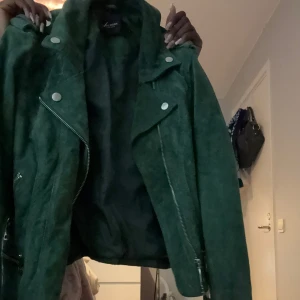 Grön mockajacka från Vero Moda - Snygg grön mockajacka från Vero Moda med coola silvriga dragkedjor och knappar. Jackan har klassisk bikerstil med flera fickor och detaljer framtill. Perfekt för dig som vill sticka ut med en färgstark look. Modellen är kortare och har lång ärm.