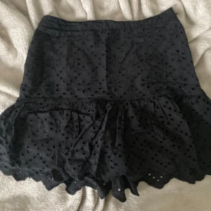 Svart broderad kjol från Lindex - Supersöt svart kortkjol från Lindex i bomull med broderade blommor och volangkant. Kjolen har en feminin och luftig känsla med fina detaljer och passar perfekt till sneakers eller sandaler. Perfekt för dig som gillar en söt och trendig stil.