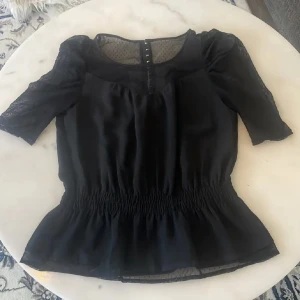 Svart blus med meshärmar, stl 36 - Säljer en svart blus med transparenta meshärmar och detaljerad mesh upptill. Blusen har smockad midja som ger en snygg siluett och knappar i ryggen. Perfekt för dig som gillar stilrena och trendiga plagg med lite extra edge.