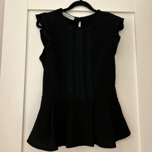 Svart volang topp från Rinascimento - Supersnygg svart topp från Rinascimento med volang nedtill och broderade detaljer längs axlarna och framtill. Ärmlös design med rund hals och liten knapp i nacken. Perfekt för dig som gillar stilrena och coola plagg med extra detaljer.