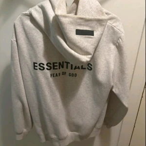 Essentials Fear of God grå hoodie - Säljer en snygg ljusgrå hoodie från Fear of God Essentials med svart tryck både fram och bak. Tröjan har huva, känguruficka och långa ärmar. Perfekt för dig som gillar streetwear och vill ha en clean look med stilren design.