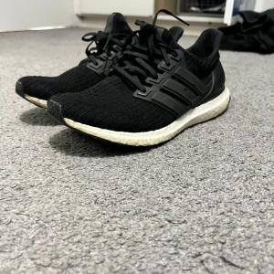 Svarta Adidas Ultraboost sneakers - Säljer ett par svarta Adidas Ultraboost sneakers med stickad ovandel och klassiska tre ränder på sidan. Skorna har vit Boost-sula för extra komfort och svart yttersula. Perfekta för dig som gillar stilrena och sportiga sneakers.