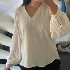 Ljusbeige blus med v-ringning från mbyM - Superfin ljusbeige blus från mbyM i storlek S. Blusen har v-ringning, långärmade puffiga ärmar med knappdetalj vid mudden och ett diskret strukturerat tyg. Perfekt till jeans eller kjol för en clean och stilren look. Blusen är i fint mycket fint skick utan defekter.
