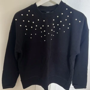 Svart sweatshirt med pärlor från Lindex - Svart sweatshirt från Lindex med coola pärldetaljer över axlar och bröst. Tröjan har rund hals och långa ärmar, perfekt för dig som gillar att sticka ut lite extra. Mjuk bomullskvalitet som är skön att ha på sig hela dagen.