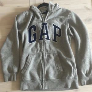 Grå hoodie från GAP med dragkedja - Säljer en grå hoodie från GAP med stor, mörkblå logga framtill. Passar som xs