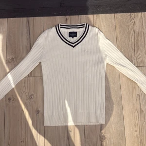 Vit ribbad tröja från Bondelid - Vit ribbstickad tröja från Bondelid med svart-vita ränder i v-ringad krage. Långärmad och med diskret broderad logga på bröstet. Klassisk och stilren modell som passar perfekt till jeans eller shorts.