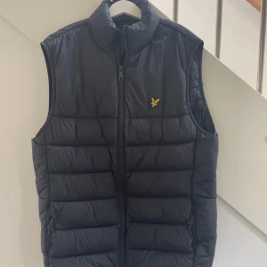  Lyle & Scott Väst - Snygg marinblå  dunväst från Lyle & Scott med gul logga på bröstet. Västen har hög krage, quiltad design och dragkedja framtill. Perfekt att slänga över en hoodie eller tröja för extra värme. Klassisk och stilren look. | Tveka inte att höra av dig vid funderingar ☺️|