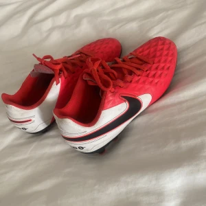 Röda Nike fotbollsskor med dobbar - Säljer ett par röda och vita Nike fotbollsskor med svart swoosh på sidan. Skorna har snörning och en strukturerad ovandel med diamantmönster för extra grepp. Yttersulan är svart med röda dobbar, perfekt för spel på gräs. Snygg design som sticker ut på planen.