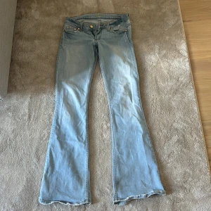 Ljusblå bootcut jeans med slitna detaljer - Säljer ett par ljusblå bootcut jeans med klassisk femficksdesign och snygga slitningar nertill. Jeansen har broderade bakfickor och låg midja. Perfekta för dig som gillar en avslappnad och retro vibe.