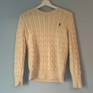 Beige kabelstickad tröja Polo Ralph Lauren - Snygg beige kabelstickad tröja från Polo Ralph Lauren med klassisk rund hals och marinblå broderad logga på bröstet. Tröjan har långärmad passform och ribbade muddar vid ärmslut och nederkant. Perfekt för dig som gillar stilren och tidlös design. Väldigt bra skick med inga defekter 
