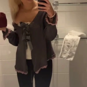 Grå stickad kofta med rosett och volang - Supersöt grå stickad kofta med offshoulder-look, stora satinrosetter framtill och ljusrosa volangkant vid ärmslut och nederkant. Koftan har lång ärm och en loose passform, perfekt för lager-på-lager och en romantisk vibe.