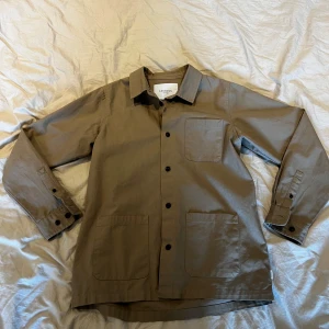 Beige overshirt från Les Deux - Stilren beige overshirt från Les Deux med klassisk krage, tre stora fickor framtill och svarta knappar. Tillverkad i bomull och har diskret broderad logga på ärmen. Perfekt lager-på-lager-plagg med relaxed vibe.