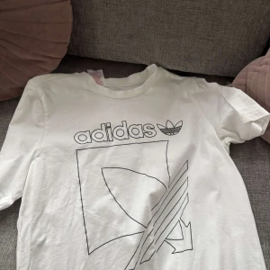 Vit t-shirt från Adidas med grafiskt tryck - Säljer en vit t-shirt från Adidas med ett stort grafiskt tryck och logga framtill. T-shirten har rund halsringning och korta ärmar. Perfekt för en sportig och avslappnad stil.