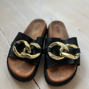 Svarta sandaler med guldfärgad kedjedetalj - Snygga svarta sandaler med bred rem i skinn och stor guldfärgad kedja framtill. Tjock sula gör dem både snygga och sköna. Stilsäkra och trendiga med chunky känsla. Philip Hog nypris 990 kr
