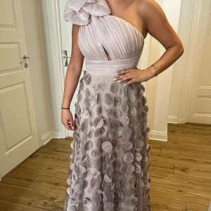 Ljus champagnefärgad långklänning - Säljer en elegant långklänning i ljus champagnefärg med en axel och stor volangdetalj upptill. Kjolen är täckt av 3D-blommor och pärlor för en unik look. Perfekt för bal eller fest! Materialet känns lyxigt och klänningen har oneshoulder-modell. Nypriset är 4000kr