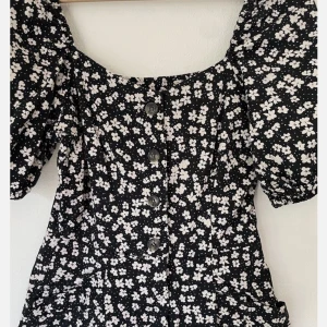 Svart blommig kortklänning med puffärmar - Supersöt svart one piece, kortklänning med skorts inbyggda, vita blommor och puffärmar. Klänningen har knappar framtill och praktiska fickor. Perfekt för sommarens alla tillfällen! Snygg och bekväm passform med markerad midja. Och två st fickor! 