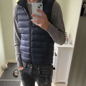 Uniqlo väst  - Snygg mörkblå dunväst med dragkedja framtill. Västen har en klassisk passform och är perfekt att bära över en tröja. Enkel och stilren design utan synliga loggor eller mönster.