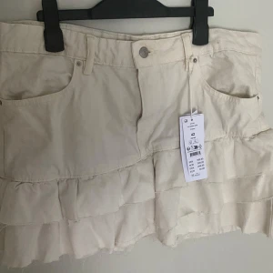 Krämvit volangkjol i denim - Säljer en krämvit kort volangkjol i denim från Gina. Kjolen har fickor fram och volanger. Perfekt att styla med sneakers eller boots för en trendig vibe. Kjolen  är helt oanvänd med lappen kvar🌸