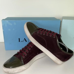 Vinröda sneakers från Lanvin med lackad tå - Säljer ett par snygga vinröda sneakers från Lanvin med ovandel i mocka och lackad mörkgrön tå. Skorna har vita sulor, vinröda skosnören och metallfärgade öljetter. Perfekta för dig som vill ha något unikt och stilrent.