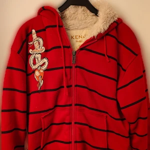 Kenji Hoodie - Röd hoodie med svarta ränder från Kenji. Jackan har dragkedja, huva med fluffigt foder och broderade motiv av orm och drake. Perfekt för dig som gillar unika detaljer och streetwear-stil. Skön, stor & varm hoodie i bra skick.  