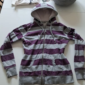 Randig hoodie från Pimkie - Mysig hoodie med breda lila och grå ränder från Pimkie. Tröjan har dragkedja, huva med fluff och två fickor framtill.