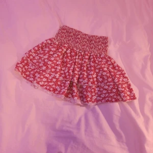 Röd blommig kortkjol med volang - Supersöt röd kortkjol med vita blommor och bred smock i midjan. Kjolen har volang och är perfekt för sommarens alla tillfällen. Lätt och luftig modell som ger en skön känsla. Stretchig i midjan för att passa alla. 