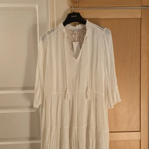 Vit långklänning med spetsdetaljer från Amisu - Vit långklänning från Amisu med vackra spetsdetaljer upptill och vid axlarna. Klänningen har trekvartsärm med volang och en lätt rynkad struktur. Perfekt för somriga tillfällen och har en romantisk känsla.