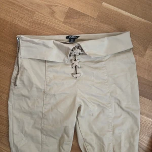 Beiga byxor med snörning och dragkedja från Murci - Säljer ett par beiga byxor från Murci med mid waist, snörning framtill och dragkedja i sidan. Byxorna har bootcut-modell och är tillverkade i ett mjukt bomullsmaterial. Finns ett litet hål som visas i 3e bilden, men går inte igenom tyget.