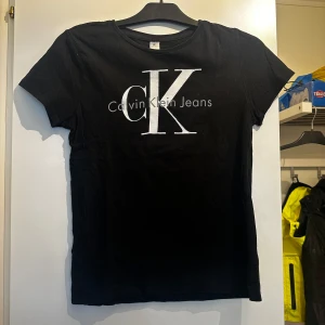 Svart t-shirt med CK-tryck - Svart t-shirt med stort vitt CK-tryck och texten Calvin Klein Jeans på bröstet. Klassisk rund halsringning och korta ärmar. Tillverkad i mjukt material som känns skönt mot huden. Aldrig använd. 