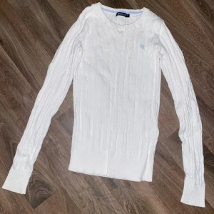 Vit kabelstickad tröja från Gina - Säljer en klassisk vit kabelstickad tröja från Gina med rund halsringning och diskret broderad logga på bröstet. Tröjan har lång ärm och ribbade muddar vid ärmslut och nederkant. Perfekt för en stilren och tidlös look.