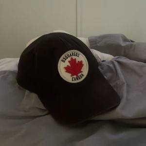 Dsquared2 keps - Svart keps från Dsquared2 med vitt runt märke framtill med röd lönnlöv och texten 'DSQUARED2 CANADA'. Justerbart spänne bak och broderad text 'DEAN & DAN CATEN' baktill. Perfekt för dig som gillar streetwear och vill sticka ut.
