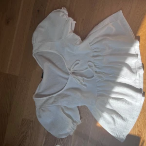 Vit topp / blus med puffärm och knyt - Supersöt vit kort blus med puffiga kortärmar och knyt i midjan. Toppen har en lätt v-ringning och är gjord i ett luftigt material som passar perfekt till varma dagar. Stilren och enkel att matcha med accessoarer. Med rosetter