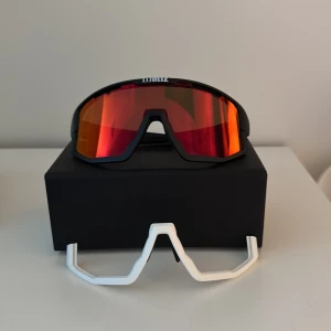 Bliz Vision sportglasögon med röd spegellins - Sportiga Bliz Vision-glasögon med matt svart båge och röd spegellins. Medföljer extra vit jawbone och näsdel. Perfekta för träning utomhus, ger bred sikt och skydd mot solen.