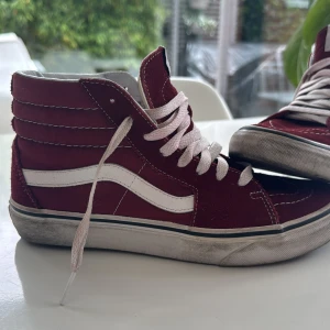 Vinröda Vans Sk8-Hi sneakers - Säljer ett par klassiska vinröda Vans Sk8-Hi sneakers med vit sula och vit sidoremsa. Skorna har högt skaft, vita snören och tydliga sömmar. Perfekta för dig som gillar streetstyle och vill ha en ikonisk look.