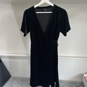 Svart omlottklänning i sammet från New Look - Säljer en svart omlottklänning i mjuk sammet från New Look. Klänningen har korta ärmar, v-ringning och knytdetalj i midjan. Perfekt för dig som gillar klassisk och stilren look.